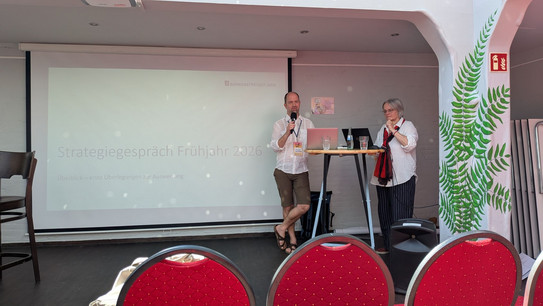 Foto: Die 2 Referent*innen stehen an einem Stehtisch, darauf ihre aufgeklappten laptops. Carsten Bender hlt ein Mikro. Im Hintergrund wird eine Präsentationsfolie an die Wand projiziert.