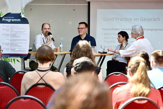Foto: 4 Referent*innen sitzen nebeneinander, die Person ganz links hält ein Mikro und neigt den Kopf Richtung Plenum, das sich im Vordergrund befindet