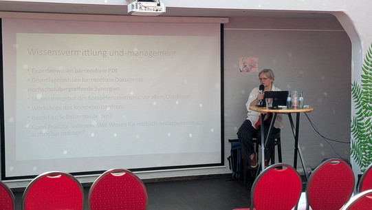 Foto: Die Referentin steht an einem Stehtisch, darauf ihr geöffneter Laptop. Sie hält ein Mikro. Im Hintergrund wird eine Präsentationsfolie an die Wand projiziert.